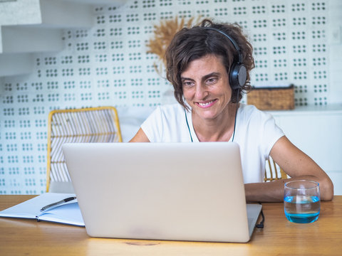 
happy smiling remote woman talking and flirting with headset, laptop, notebook, pen and glass in casual outfit sitting on a work desk in her living room in her home office having video chat
