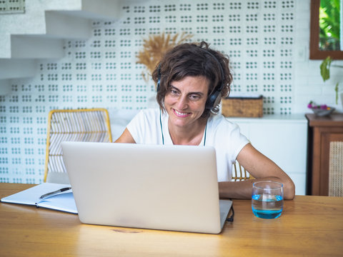 
happy smiling remote woman talking and flirting with headset, laptop, notebook, pen and glass in casual outfit sitting on a work desk in her living room in her home office having video chat
