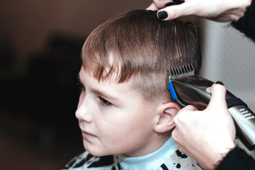 Fototapeta premium Toned image.Hairdresser makes a stylish hairstyle. The woman is standing and making haircut for blonde boy. Close up of hands.