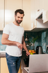 Selective focus of smiling man looking at laptop while cooking in kitchen