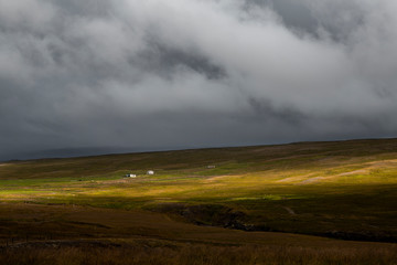 landscape in iceland. There is a lonely house in the middle of a green meadow. the sky is full of clouds and we can see torrents of water falling in the middle of the sun's rays