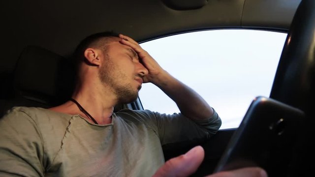 A man in a car rubs his head with his hand and looks into his smartphone. He's driving, he's in a bad mood. Thinks and shakes his head. A close-up. Hd.