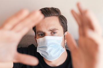 Young handsome man in medical mask sitting at home during isolation time. Looking seriously at the camera, putting his hands forward the camera