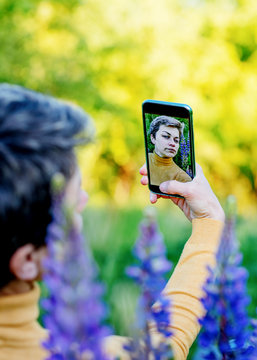 Woman taking photos of purple Lupines on the phone on the green grass at sunset