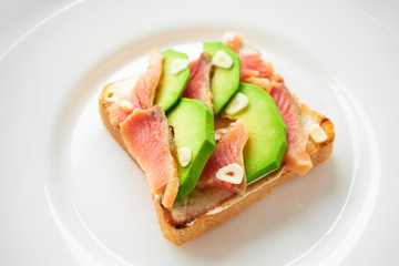 Sandwich with salmon, avocado, white sauce and garlic slices on toasted bread on a white plate