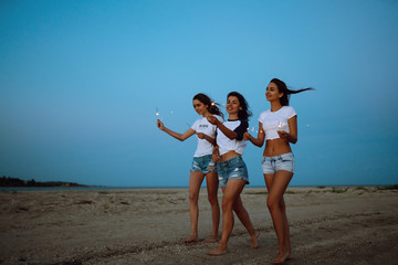 Three beautiful girls celebrating, holding sparklers  on the beach at night. Young teenagers enjoying on beach holiday. Summer holidays, vacation, relax and lifestyle concept.