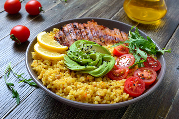 Bulgur with grilled chicken breast, fresh vegetables, avocado, arugula. Diet lunch on a wooden table. Healthy food.