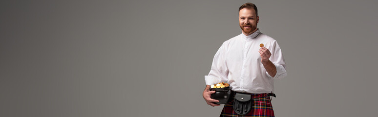 smiling Scottish redhead man in red kilt holding potty with gold coins on grey background,...
