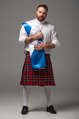 Scottish redhead man in red kilt with flag of Scotland on grey background