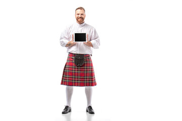 smiling Scottish redhead man in red kilt presenting digital tablet on white background
