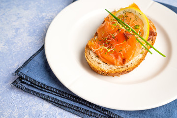 Norwegian smoked salmon with lemon slice, chives and sprouts decoration on a white plate with blue textured backdrop. Food photography with sidelight.