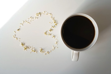 On a white background is a coffee mug and a heart of white flower petals is laid out next to it.