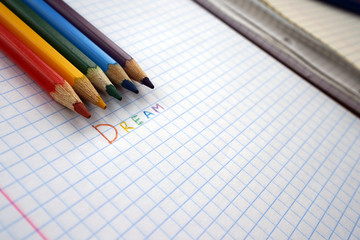 The inscription DREAM written in colored pencils, next to a checkered sheet of paper lay pencils