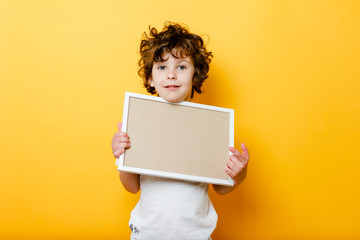 curly cute boy in white T-shirt holds an empty frame for text, smiling and shows up finger on bright yellow background.white signboard or banner - copy space concept to fill text. Message board