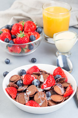 Trendy small chocolate cereal pancakes served with condensed milk, strawberry, blueberry and orange juice, in white bowl, vertical