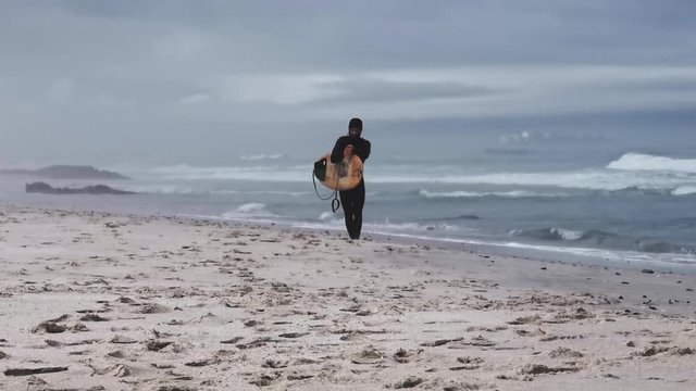 A surfer after leaving the water walks along the beach on a misty, hazy dawn