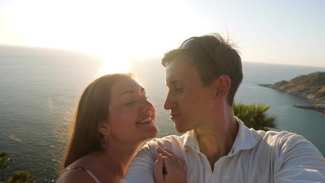 couple makes selfie and lady in summer dress hugs and kisses guy against endless blue ocean under bright back sunlight closeup