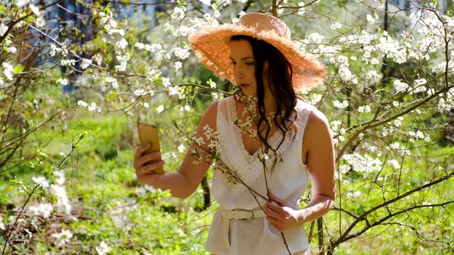 brunette woman with long hair in a straw hat stands in the apple orchard and looks at the camera