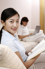 Woman reading book, man using laptop in the background