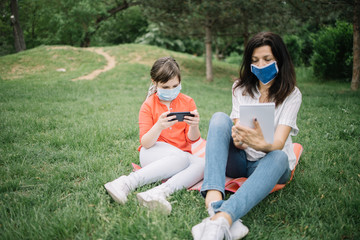 Woman wearing antivirus mask while resting in park with her daughter. Little girl with antivirus mask using phone while lying on mat in forest with her mother who is holding notebook.