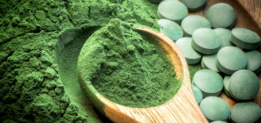 Green algae in powder and pills - chlorella, spirulina in wooden spoon on wooden background - closeup - banner design.