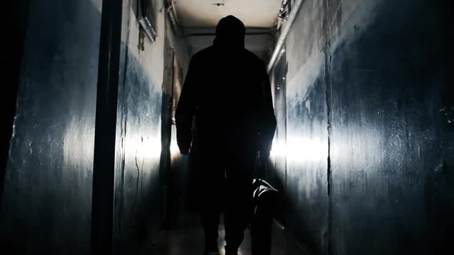 Silhouette of Unrecognizable Criminal Man Wearing Black Jacket and Carrying a Bag Walking Away From the Camera Along Hallway in Old Apartment Building Long Dark Hallway