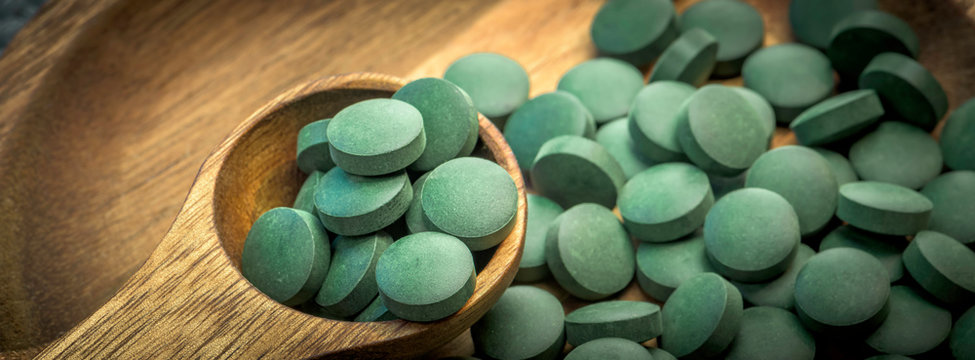 Green algae in tablets - chlorella, spirulina in wooden spoon on wooden background - closeup banner design.