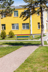 Fototapeta na wymiar Rainbow colored bench on the background of yellow building facade. Bright colorful street photography, multicolor exterior architecture design. Summer sunny day, empty park with no people.