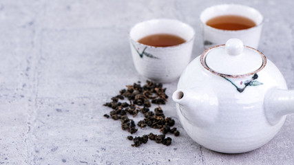 Hot tea in white teapot and cups on a sieve over bright gray cement background, closeup, copy space design concept.