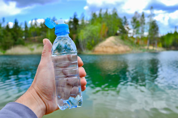 A man's hand holds a water bottle close-up against the background of clear water of the lake with a...