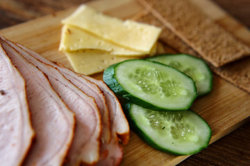 Slicing ham, cucumbers and cheese with ruddy thin breads lie on a wooden board.