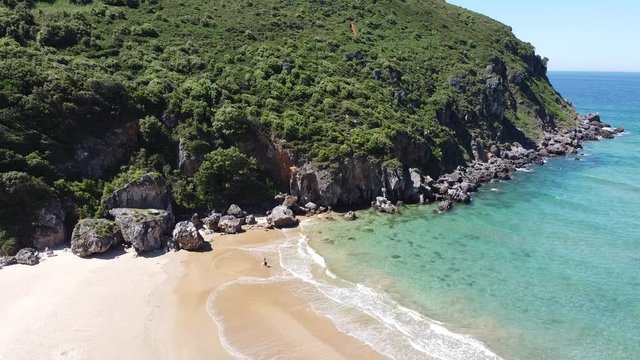 Imagen a&eacute;rea est&aacute;tica desde mucha altura de una playa, peque&ntilde;as olas rompen suavemente mientras una persona se acerca al agua para refrescarse, una monta&ntilde;a con vegetaci&oacute;n frondosa preside la escena