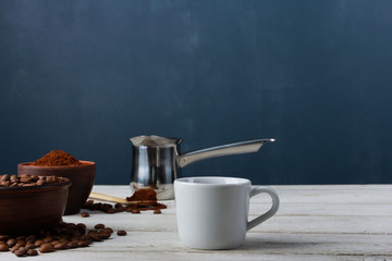 White coffee cup, Arabica beans in clay bowls, ground powder, metal Turkish pot on while table against dark blue wall. Side view, copy space. Coffee shop, morning, baristas workplace concept