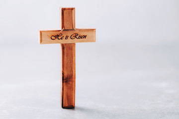 Wooden cross with text He is risen on grey background. Reminder of Jesus sacrifice and Christ resurrection. Easter passover. Eucharist concept. Christianity symbol and faith