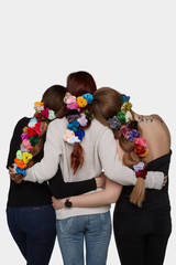 Back view of three girls with colorful velvet scrunchies in their long braids. The blonde, brunette and ginger lady are wearing jeans and hugging on the white background. 