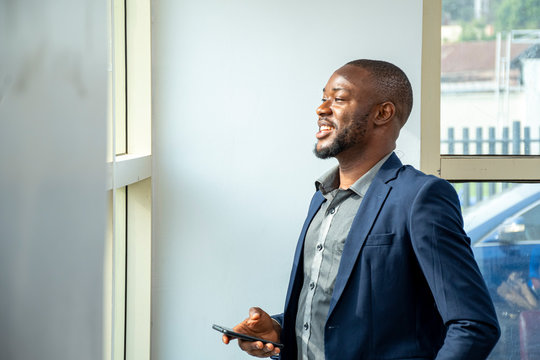 Young African Business Man Wearing A Suit Smiling And Looking Out The Window In His Office