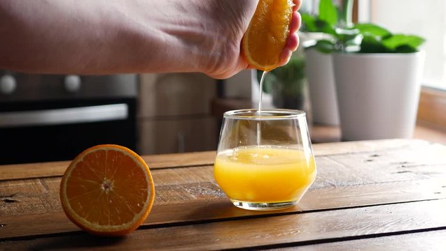 Hand squeezing out fresh orange into glass. Citrus juice drains from the pulp and drips. On kitchen background.