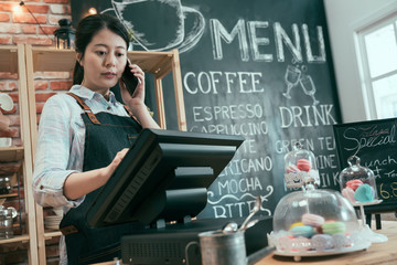 young asian japanese woman staff working in cafe store. lady waitress talking on cellphone while customer taking order. female barista using point of sale terminal and mobile phone in coffee shop. - Powered by Adobe