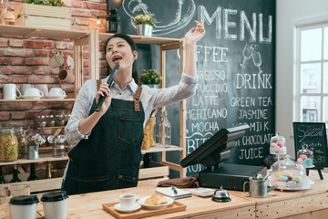 happy woman barista posing in cafe bar counter working emotional listening music singing screaming with pastry tongs as microphone. young chinese girl enjoy dancing and having fun in coffeehouse