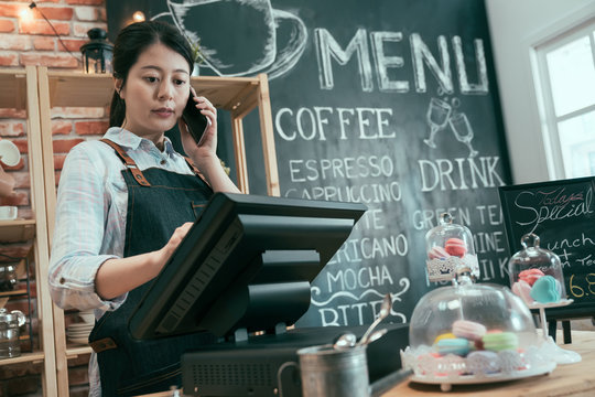 young asian japanese woman staff working in cafe store. lady waitress talking on cellphone while customer taking order. female barista using point of sale terminal and mobile phone in coffee shop.