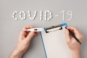 COVID-19 flu epidemic. The inscription Covid-19 of pills, tablets, notepad and a thermometer in men's hand on a gray background, Flat lay, copy space. Temperature measurment