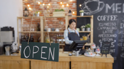 Smiling waitress standing at counter busy working in morning using pos machine. slate blackboard showing with open sign in cafe bar. beautiful waitress barista in back blur prepared making coffee