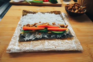Preparing vegan homemade sushi and rolls on wooden board. View of process of preparing rolling sushi. Sushi with slice of vegan tofu, sticky white rice, avocado, carrot, spring onion.