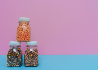 Small bottles of dry cereals on pink and blue background. Different types of cereals and legumes, close up. Set of whole grains - buckwheat, red lentil, beans, peas, red beans. Copy space.