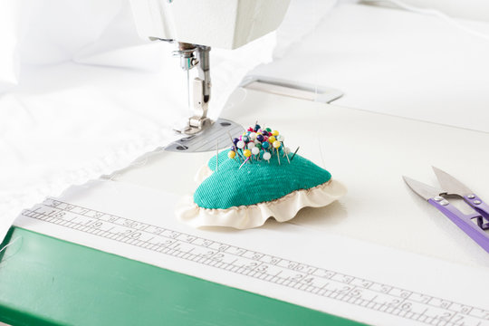 The mechanism of the sewing machine sews fabric and a pillow for pins with needles, scissors on a white with a green table. Workplace sewing master, sewing studio, apprentice