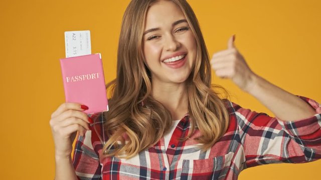 Young happy girl isolated over yellow wall background holding passport with tickets pointing