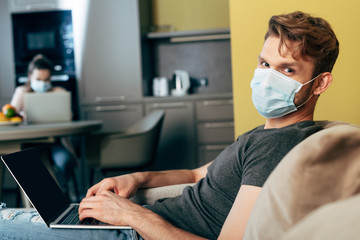 selective focus of freelancer in medical mask using laptop with blank screen near girlfriend