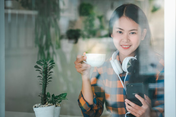 A beautiful Asian woman in a coffee shop is happy to eat coffee and order online via mobile phone. Charcoal image through a glass in a coffee shop The concept of an Asian woman in a coffee shop