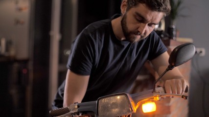 Man repairs a blue scooter or motorcycle in home brick garage. Man checks the headlights of a motorcycle. Self-isolation due to coronavirus. Sports moped or bike stands in a home brick garage.
