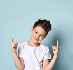 teenage kid in white t-shirt. He smiling, raised his forefingers up, posing against blue studio...